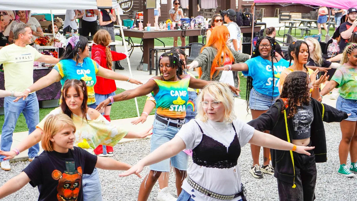 A large group of dancers of all ages stand with arms outstretched mid-choreography outdoors, several wearing matching tie-dye camp shirts, at Knox Pop Con.